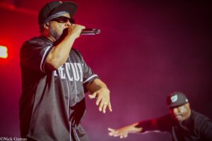 Male rapper wearing a black cap and sunglasses sings into a microphone on a red-lit stage.