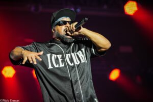 Male rapper performing on stage, microphone in hand, wearing sunglasses and a black cap, under red stage lights.