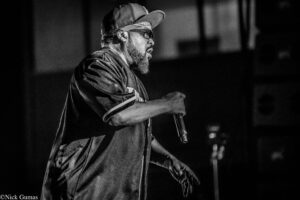 Black-and-white photo of a bearded rapper on stage, wearing a cap, sunglasses, and a bandana, holding a microphone.