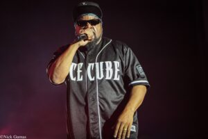 Hip-hop performer on stage wearing sunglasses and a black cap, rapping into a microphone with a black jersey that reads ICE CUBE on the chest.