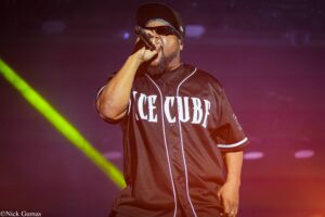 Male rapper on stage, wearing a black baseball jersey and sunglasses, singing into a microphone under colored stage lights.