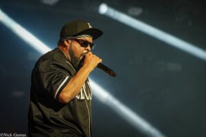 Male singer wearing a black cap, sunglasses, and bandana, singing into a microphone on a dimly lit stage with bright beams behind him.