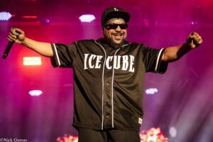 Hip-hop artist on stage with a black Ice Cube jersey, arms spread, microphone in hand, colorful pink/purple lights in the background.