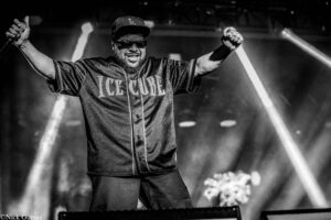 Ice Cube on stage with arms raised, holding a microphone, wearing a LA cap and a jersey that reads ICE CUBE (black-and-white photo).