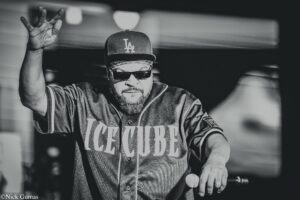 Rapper Ice Cube performing on stage, wearing a Los Angeles cap, sunglasses, and a jersey, raising his left hand.