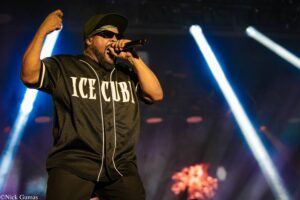 Male rapper on stage wearing a black 'ICE CUBE' jersey, sunglasses and cap, singing into a handheld mic with a raised fist, blue stage lights in the background.