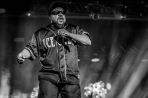 Male hip-hop performer wearing a cap and sunglasses, singing into a microphone on stage in a smoky setting (black and white).