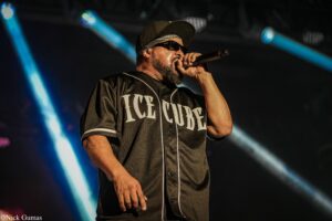 Male performer wearing a black cap, sunglasses, and a dark jersey with large white lettering, singing into a handheld microphone on stage with blue spotlights behind him.
