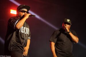 Two male performers on stage singing into microphones under red and white stage lights, wearing black clothing and sunglasses (left) and cap (right)