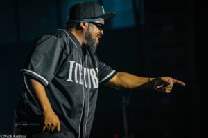 Male performer on stage, wearing a black cap, sunglasses, and a dark jersey, holding a microphone and extending an arm forward.