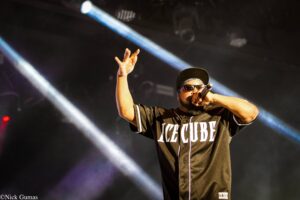 Hip-hop artist wearing a black Ice Cube jersey, singing into a mic with one arm raised on stage under blue spotlights.