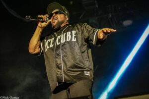 Male rapper performing on stage, wearing a black Ice Cube jersey, sunglasses and cap, holding a microphone and gesturing with his other arm amid stage lights.