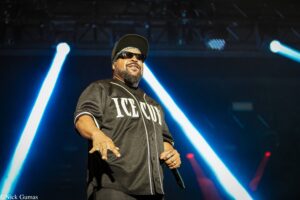 Hip-hop artist Ice Cube performing on stage in sunglasses and cap, microphone in hand amid blue stage lights.