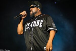 Male performer wearing a black baseball jersey with white piping, sunglasses, and a cap, singing into a handheld microphone on a dark stage.
