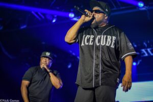 Male rapper on stage wearing a black baseball jersey with 'ICE CUBE' across the chest, microphone in hand, blue stage lights in the background.