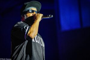 Male singer on stage, wearing a cap and sunglasses, singing into a handheld microphone with blue stage lights behind him.