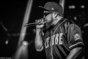 Black-and-white photo of a male rapper wearing a cap and sunglasses, singing into a microphone on stage during a live show.