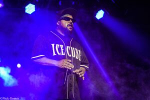 Male performer on a smoky blue-lit stage wearing a baseball cap and sunglasses, holding a microphone mid-song.