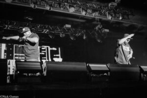 Two male performers on a dimly lit stage; left artist raps into a microphone with an outstretched arm, right artist raises a hand to the crowd (black and white photo).