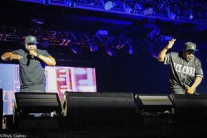 Two male performers on a dimly lit stage with blue lights; one sings into a microphone, the other raises a hand, wearing an Ice Cube jersey and LA cap.