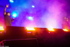 Two performers on a smoky stage under blue, purple, and pink lights; left singer raises a hand and holds a microphone.