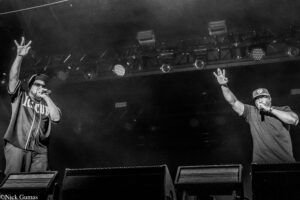 Two male performers on a dark stage; left rapper raises three fingers while singing into a mic, right rapper shows a V-sign with a headset mic on.
