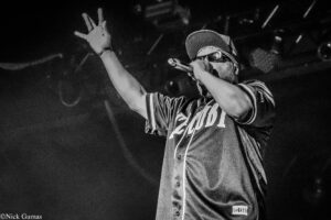 Rapper on stage in grayscale, wearing cap and sunglasses, microphone in hand with one arm raised.