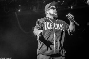 Male performer on stage wearing a cap and sunglasses, holding a microphone.