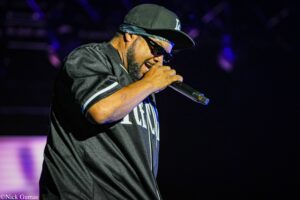 Male performer wearing an LA cap and sunglasses sings into a microphone on a dark stage.