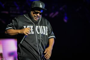 Male performer wearing a black Los Angeles cap, sunglasses, and a dark jersey, holding a microphone on stage with purple lighting in the background.
