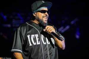 Male performer on stage, wearing a black cap, sunglasses, bandana, and a black jersey, smiling while holding a microphone.