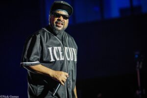 Male performer on a dark stage, wearing a black cap, sunglasses, and a black jersey that reads LEGEND, while holding a microphone.
