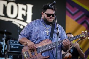 Musician wearing sunglasses and a patterned shirt playing a wooden bass guitar on stage, with a microphone in front and colorful backdrop behind him.