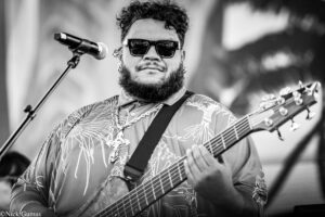 Musician wearing sunglasses and a patterned shirt plays a guitar on stage, singing into a microphone.