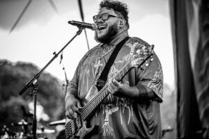 Musician singing into a microphone while playing a bass guitar on an outdoor stage (black and white)