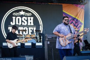 Live band on an outdoor stage: guitarist in black left, bassist/vocalist in a purple shirt center-right singing into a microphone, drummer in back, colorful backdrop and large screen behind them.