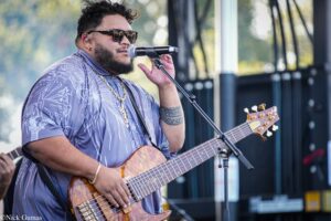 Male guitarist singing into a microphone on an outdoor stage, wearing sunglasses and a purple patterned shirt with a gold chain.