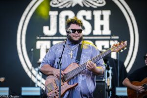 Musician with beard and sunglasses playing a wooden bass guitar on stage during a live concert.