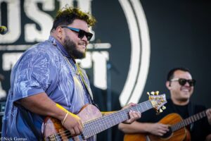 Two guitarists perform on stage: foreground man in sunglasses and a patterned blue shirt, playing an acoustic guitar; a second guitarist smiles in the background near a mic stand.