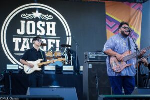 Two musicians perform on a stage: a guitarist in black with a cream guitar on the left, and a bearded singer with sunglasses and a bass guitar on the right, in front of a large circular logo that reads 'JUST TAKE' on the backdrop.