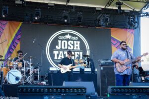Band performs on an outdoor stage: drummer on left, guitarist center, and bassist right, with a large circular 'JOSH' logo on a screen behind them.