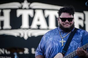 Musician wearing sunglasses and a blue patterned shirt plays an electric guitar on stage with a blurred backdrop behind him.