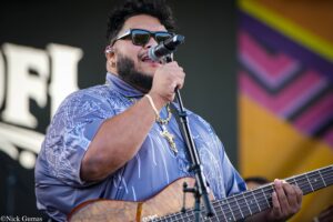 Male singer wearing sunglasses and a patterned blue shirt, singing into a microphone while playing a wooden guitar on stage.
