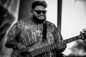 Black-and-white close-up of a bearded musician wearing sunglasses and a patterned shirt, playing a long-neck bass guitar on stage