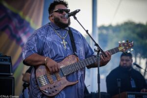 Musician on stage singing into a microphone while playing a brown bass guitar, wearing sunglasses and a gold cross necklace outdoors.
