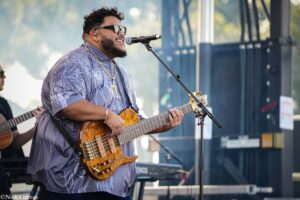 Musician wearing sunglasses plays a wood guitar at an outdoor stage, singing into a microphone. (Informative alt text)