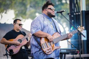 Male musician wearing sunglasses and a patterned blue shirt sings into a microphone while playing a wooden electric guitar at an outdoor concert; another guitarist is visible in the background.