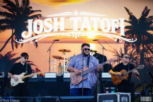 Live outdoor concert with three guitarists on stage during sunset, large tropical backdrop and palm silhouettes behind them.