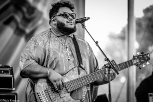 Male bassist performing on stage, wearing sunglasses and a patterned shirt, gripping a wooden bass guitar near the neck with a microphone in front, in black-and-white.