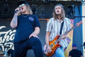 Two male rock musicians perform on stage: vocalist left with microphone, cap, sunglasses, and tattoos, and guitarist right in a striped shirt with a sunburst electric guitar.